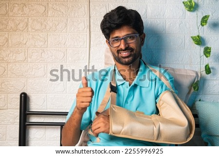 happy smiling young man with broken hand at bedroom showing thumbs up gesture by looming at camera - concept of recovered, successful medical treatment and rehabilitation