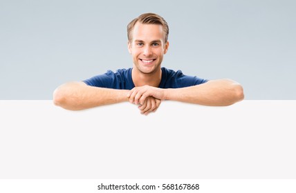 Happy Smiling Young Man Showing Blank Signboard With Empty Copyspace Area For Slogan Or Advertising Text Message, Over Grey Background. Caucasian Male Model In Concept Studio Shoot.