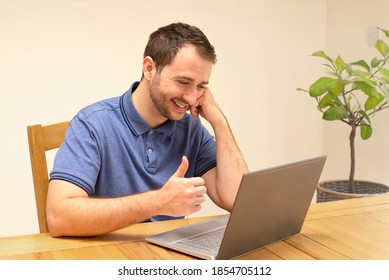 Happy Smiling Young Man Sitting At Table Indoors At Home Using Laptop Computer Working Studying. Talking To Someone On Computer Showing Thumbs Up.  Communication Online. 