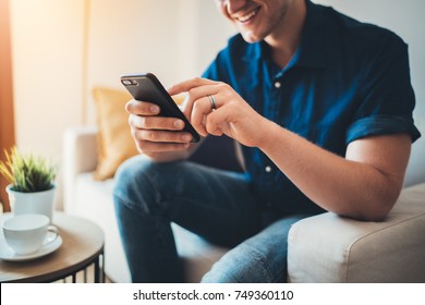 Happy lächelnden jungen Mann mit modernem Smartphone-Gerät, während er zu Hause auf Sofa sitzt, moderne Design-Inneneinrichtung, fröhlicher hipster-Typ, der eine SMS-Nachricht in sozialen Netzwerken tippt