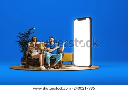 Happy smiling young man and woman, couple sitting couch near giant 3D model of mobile phone with empty screen over blue background. Shopping, presents, sales. Mockup for text, ad, design, logo