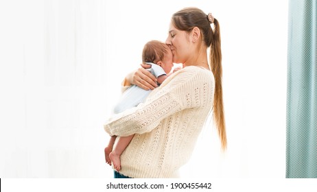 Happy Smiling Young Mother Cuddling And Kissing Her Newborn Baby Boy Against Bright Sun Shining Through Window. Concept Of Family Happiness And Loving Parents With Little Children.