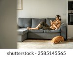 Happy smiling young student woman lying on couch, listening music in headphones on mobile phone application. Relaxing girl at new real estate apartment home using mobile phone online app. Copy space