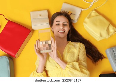 Happy smiling young woman with eyeshadows palette and handbags lying on yellow background - Powered by Shutterstock