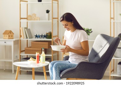 Happy smiling young woman having fresh takeaway lunch from food container sitting in an armchair in sunny living-room at home. Healthy eating, ordering good quality food online, meal delivery service