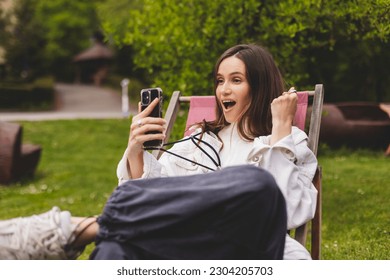 Happy smiling young woman hold smartphone with excited face expression, unable to believe that she bet played, win in lottery, read good news, can't believe in her fortune. Girl sitting in park. - Shutterstock ID 2304205703