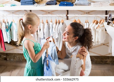 happy smiling young woman with little daughter selecting blue kids apparel in clothes store

