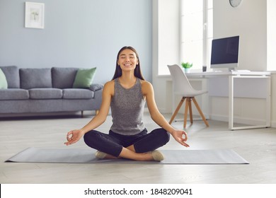 Happy Smiling Young Woman Practicing Meditation In Easy Pose Taking Break From Work In Home Office. Positive Asian Girl Sitting Legs Crossed On Yoga Mat Reaching Zen, Enjoying Stress Relief Exercise