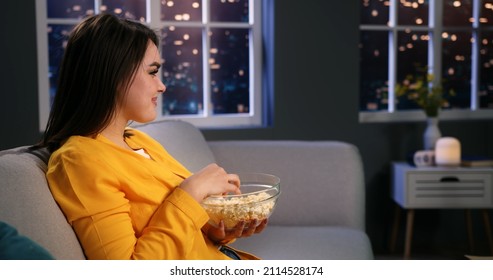 Happy Smiling Young Woman Sitting On Couch At Home Watching Funny Tv Programme Comedy Film, Eating Popcorn And Laughing. Side View Close Up.