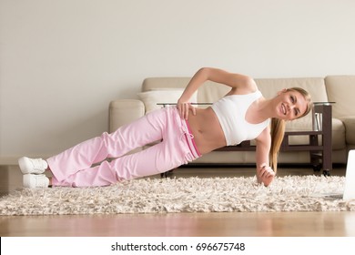 Happy Smiling Young Woman In Sportswear Doing Side Plank While Lying On Floor On Carpet At Living Room. Lady Watching Fitness Online Videos On Laptop And Tries To Get Flat Stomach With Abs Exercises