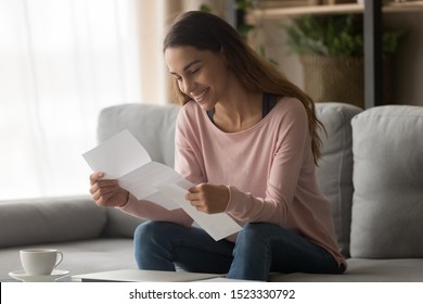 Happy Smiling Young Woman Student Customer Holding Paper Bank Statement Open Read Mail Letter With Good News, Loan Approval, Receive Scholarship Admission Money Tax Refund Concept Sit On Sofa At Home