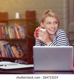 Happy Smiling Young Woman Taking A Coffee Break As She Sits At Her Laptop Computer Working From A Home Office With Bookcase