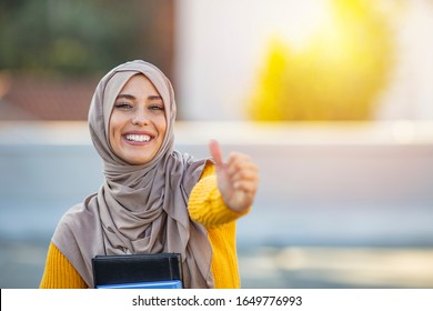 Happy smilling Muslim woman student with books showing thumbs up gesture. Muslim woman giving thumbs up sign. Beautiful female middle eastern college student in front of campus.