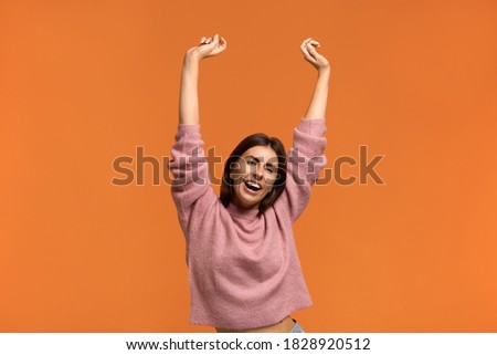 Happy sound.Energized happy woman in pink knitted sweater raises hands joyfully, being in high spirit, jumps playfully, dances to favourite music. Isolated on orange background
