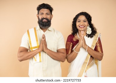 Happy South indian couple wearing traditional white dress standing in a greeting pose to Namaste hands isolated on beige background, man wearing lungi and woman wearing saree.