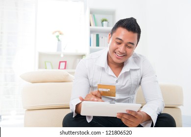 Happy Southeast Asian Man Online Shopping, Hands Holding Credit Card And Computer Tab Sitting At Home.