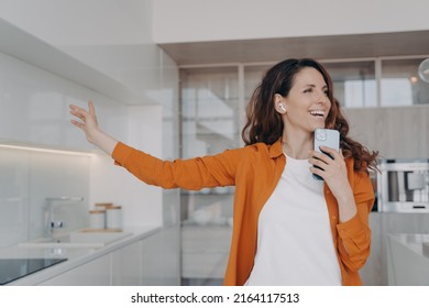 Happy Spanish Girl In Airpods Is Singing With Mobile Telephone As With Microphone. Woman Having Fun, Relaxing And Dancing. Application For Music Listening Online. Housewife Is Singing Song At Home.