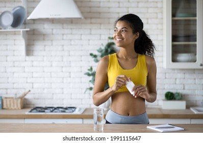 Happy sportive african american woman in sportswear holding a bottle of nutritional supplements, looking happily out the window, healthy lifestyle