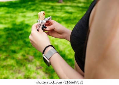 Happy Sportive Woman Wearing Sports Bra Standing On City Park, Outdoors Opening Protein Bar Package. Eating Protein Bar, Healthy Food Or Snacks. Green, Sunny Day And Sportive Concepts.