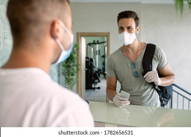 Happy Sportsman Wearing Protective Face Mask While Checking Into Health Club And Talking To Receptionist. 