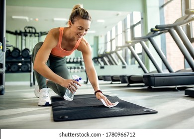 Happy sportswoman cleaning exercise mat with disinfectant after sports training in health club. 