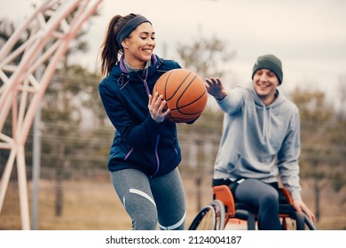 Happy Sportswoman And Her Friend In Wheelchair Having Fun Together While Playing Basketball Outdoors.