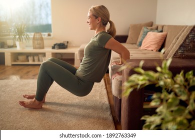 Happy sportswoman leaning on the sofa and practicing triceps dips during home workout.