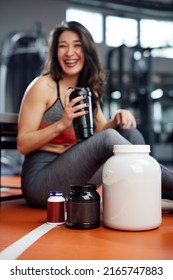 A Happy Sportswoman Sits On A Gym Floor And Drinks Protein Drink. Selective Focus On Jars And Bottles With Protein Powder, Supplements And Pills.