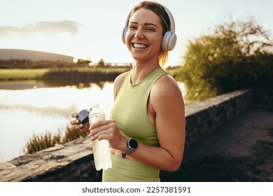 Happy sportswoman taking a water break outdoors. Fit woman standing next to a lake and looking at the camera. Woman working out with headphones in the morning. - Powered by Shutterstock