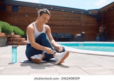 Happy sportswoman tying shoelace on her sneakers while exercising by the pool in her backyard. Copy space. - Powered by Shutterstock