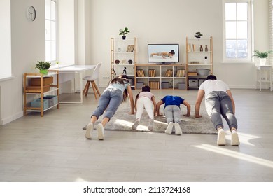 Happy Sporty Family Doing Physical Exercises At Home. Mum, Dad And Kids Practising Push Ups Together While Watching A Video Workout Lesson With A Gym Fitness Instructor On The TV Screen, Back View