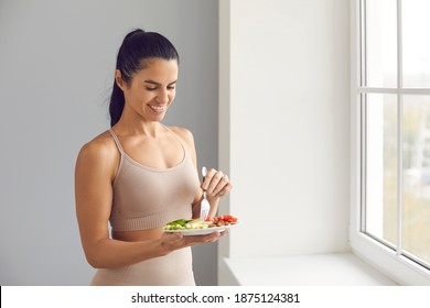 Happy Sporty Woman Having Meal And Enjoying Food. Smiling Fit Female Standing By The Window At Home, Holding Plate And Eating Delicious Vegetarian Lunch Or Breakfast. Sticking To Healthy Diet Concept