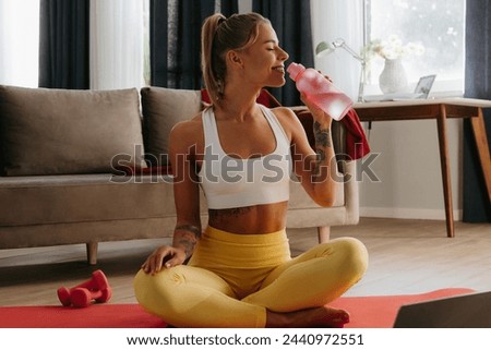 Happy sporty woman holding water bottle while resting on exercise mat at home with laptop on foreground