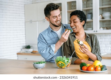 Happy spouses in love together in the kitchen,caucasian man feeding vegetables to his beloved woman, they spend time together, smile. Healthy eating, take care of health - Powered by Shutterstock