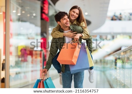 Happy Spouses Shopping And Having Fun, Husband Carrying Wife Piggyback And Holding Colorful Shopper Bags In Modern Hypermarket On Weekend. Great Seasonal Sales And Commerce Concept