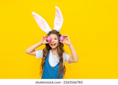 Happy spring Easter. The little girl closes her eyes with eggs and smiles broadly. A charming child with rabbit ears in a blue sundress on a yellow isolated background. - Powered by Shutterstock