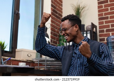 Happy Startup Owner Celebrating Bussiness Achievement Sitting At Desk Doing Success Hand Gesture In Red Brick Office. Entrepreneur With Glasses Enjoying Turnover Results And High Sales Numbers.