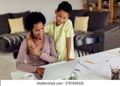 Happy Stay At Home Mother Working On A Computer While Her Son Is Embracing Her And Looking At What's She Doing. 