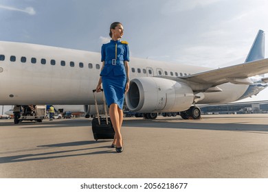 Happy stewardess holding black suitcase and walking on runway with airplane in the background