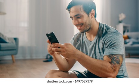 Happy Strong Athletic Fit Man in T-shirt and Shorts is Using a Mobile Phone After Morning Exercises at Home in His Spacious and Bright Living Room with Minimalistic Interior. - Powered by Shutterstock