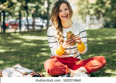  Happy Student Female Sitting On The Green Grass At The College Campus On A Sunny Day, Have Lunch And Studying Outdoors. A Smiling Young Woman Takes A Rest Eating Fast Food And Learning In The Park.