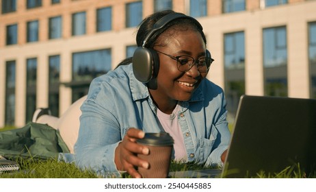 Happy student girl African American woman on lawn lying on green grass with wireless computer listening music in headphones study with laptop studying distant in park outdoors drinking takeaway coffee - Powered by Shutterstock