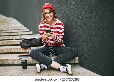 Happy student girl in earphones sitting outside with smartphone in hands having coffee break after classes, looking at camera with toothy beaming smile, dressed in striped funny outfit and glasses