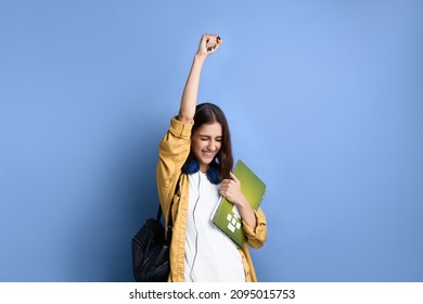 Happy student girl says yes, passed an important test, raises hand with fist in excitement, smiles sincerely, holding books, wearing yellow shirt, white t-shirt, black bag and headphones over neck - Shutterstock ID 2095015753