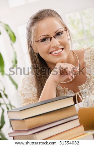 Happy student girl sitting at desk with pile of books, looking at camera, smiling.