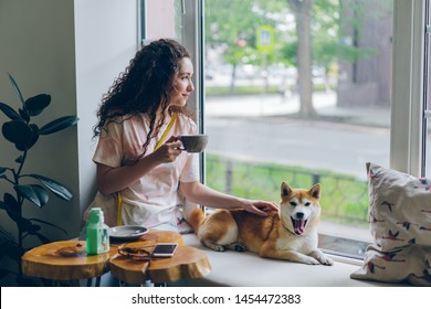 Happy student pretty girl is stroking adorable shiba inu dog sitting in cafe on window sill with cup of tea and enjoying leisure time. People and relaxation concept.