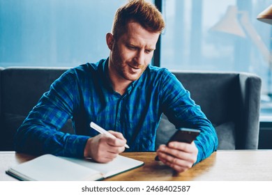 Happy student with red hair found important information on website for coursework.Cheerful young man writing down text in copybook reading incoming notification on modern smartphone.  sport bet win - Powered by Shutterstock