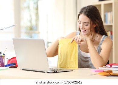 Happy student woman with laptop opening padded envelope sitting on a desk at home
