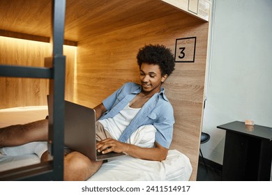 happy and stylish african american student networking on laptop on double-decker bed in hostel