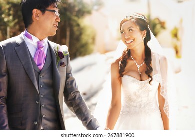 happy stylish asian bride and groom look at each other and holding hands  on street of island Santorini 